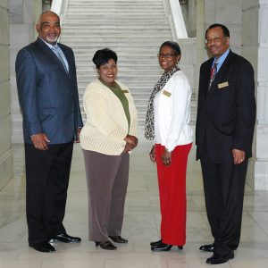 African-American men in suits and African-American women in white sweaters smiling in hallway with stairs behind them