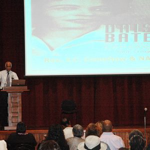 African-American man with gray hair speaking at lectern on stage with screen behind him