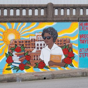 African-American girl holding "Black History" book with stack of books on her left side and roses with multistory school building behind her painted on concrete wall