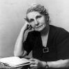 Older white woman in black posing with an open book on a table