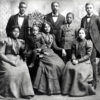 African-American women in formal dresses with a child sitting in chairs while men in suits stand behind them