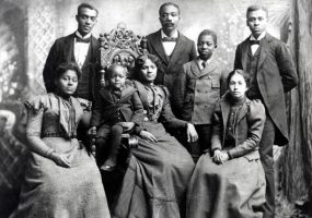 African-American women in formal dresses with a child sitting in chairs while men in suits stand behind them