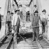 White men with hats and tools and push cart on steel truss bridge