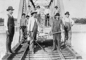 White men with hats and tools and push cart on steel truss bridge