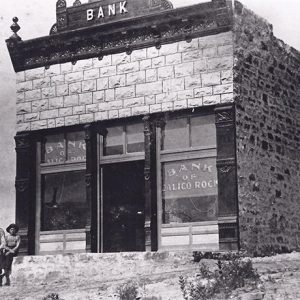 Single-story brick building with plate windows with three white people in hats sitting in front