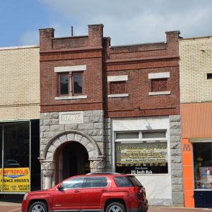 Two-story brick bank and storefront buildings on street with red SUV parked in front of them