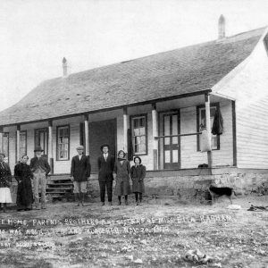 White men women and girls standing outside single-story house with covered porch and slanted roof