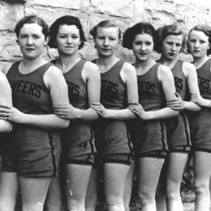 Group of young white women in sleeveless shirts and shorts standing in line against a brick wall with ball