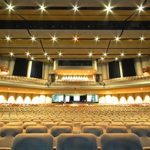 Interior of theater looking up over rows of seats to the balcony with bright lights and dark curtains