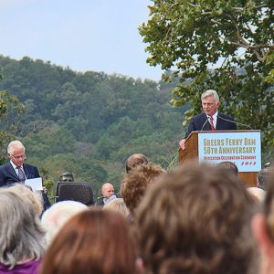 White man in suit speaking into microphone at lectern to crowd with white man and African-American man waiting on stage