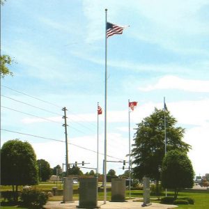 Engraved stone monuments arranged in circle formation with flag poles in the center with trees and traffic lights in the background