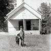 Young white child in overalls with dog standing in front of single-story building with two front doors