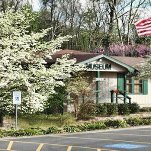 Single-story building with covered porch blooming trees and flag pole in front yard taken from parking lot