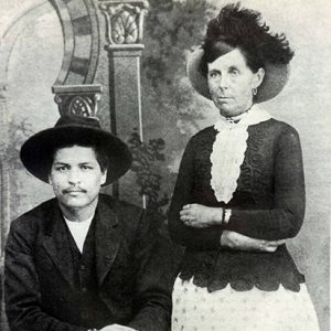 Cherokee man in hat and suit sitting next to white woman standing in hat and dress with her arms crossed