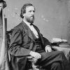 White man with beard in suit sitting in chair with stack of books on table next to him