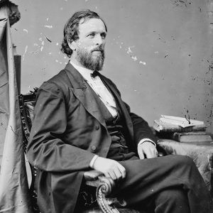 White man with beard in suit sitting in chair with stack of books on table next to him