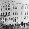 Crowd of people on steps of three-story building with parked cars