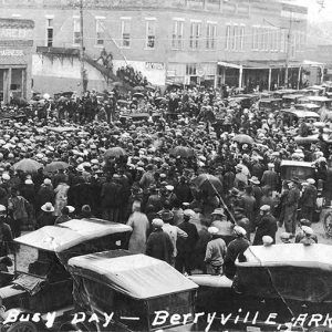 Large crowd of people and cars in street between rows of multistory brick buildings