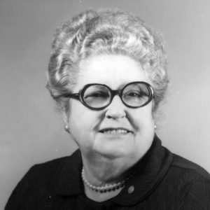 Older white woman with wavy pulled up hair and round glasses wearing a dark top and pearls