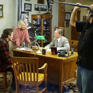 White woman talking to young white man and older white man in office on film set with white boom microphone operator in the foreground