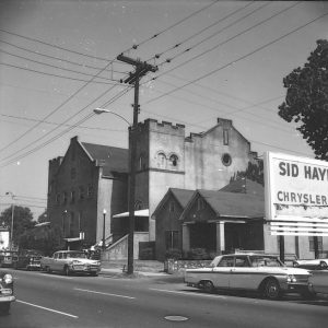 Multistory church and house on street with parked cars and power lines in the foreground