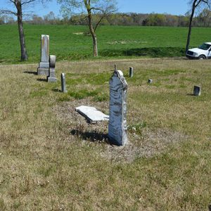 Stone monuments and gravestones in rural cemetery with green field next to it and car on driveway