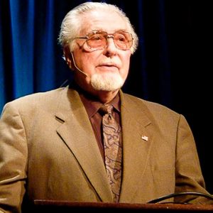 white man with white hair and goatee and tinted glass in brown suit speaking at lectern labeled "Ozark UFO Conference, Eureka Springs, Arkansas" in front of blue curtain