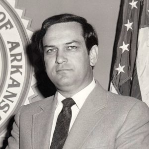 White man in suit standing with State Seal and flag behind him