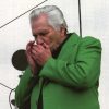 White man with gray hair in green jacket and black shirt playing harmonica