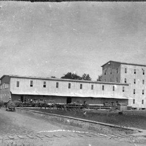 Wagons loaded with rice outside multistory mill buildings with smoke stack