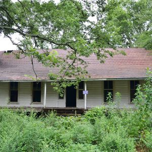 Abandoned single-story house with angled roof and covered porch in overgrown yard with trees