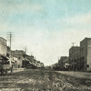 Horse drawn wagon on dirt road with multistory brick storefronts and telephone poles on both sides of it