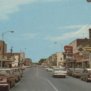 Multistory brick storefronts with signs and traffic on two-lane road lined with cars