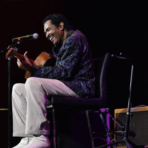 Older African-American man with mustache sitting with guitar on stage singing into a microphone