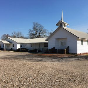 Single-story church building with cupola steeple and wing with covered porch on gravel parking lot