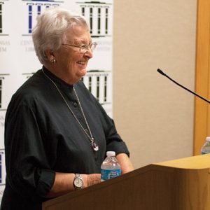 Older white woman in black shirt and long necklace speaking at lectern