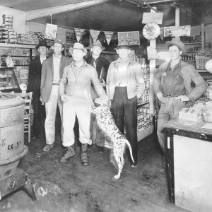 Group of white men wearing hats inside a store with a cast iron stove and Dalmatian in the foreground