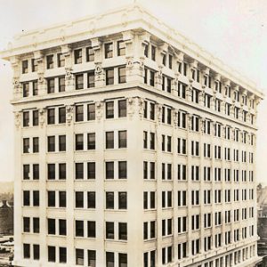 Tall multistory building on city street corner with horse-drawn carriages and people