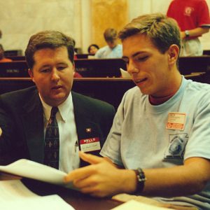 White man in suit sitting at desk in House chamber talking to young white man