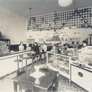 two people sitting at the counter and one person standing behind it inside a restaurant with display cases