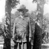 Old white woman in military-style uniform with hat standing in between two trees