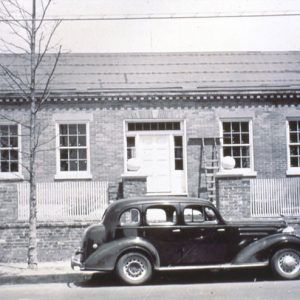 Single-story brick house inside fence with brick columns and parked cars on street outside it