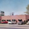 Brick building with oval shaped windows with cars in parking lot and hanging "Little Italy" sign on two lane street