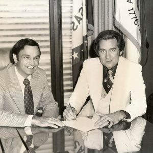 Two white men in suits sitting at desk in office with flags and telephone on a table behind them