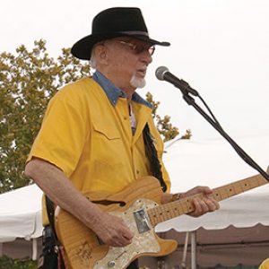 Older white man in a yellow shirt and black hat playing an electric guitar on stage in front of a microphone