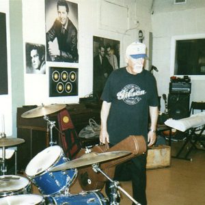 Older white man with ball cap and guitar case standing in recording studio with drum set and photographs on the walls