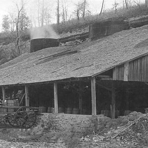 White man sitting on wagon loaded with barrels parked outside wooden pavilion with smoke stack