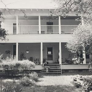 White man sitting on steps of two-story house with covered porch and balcony