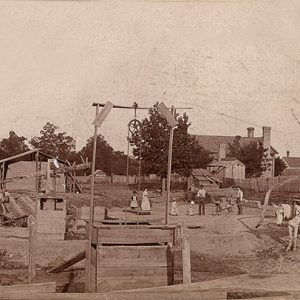 Men, woman and children in brickyard with horse drawn carts and buildings in the background