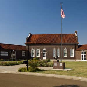 Multistory brick building with single-story wings on grass with flag pole and bell on brick pedestal next to paved driveway